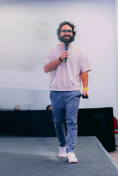 Euriler Jubé sorrindo no palco, de camiseta clara, com microfone na mão